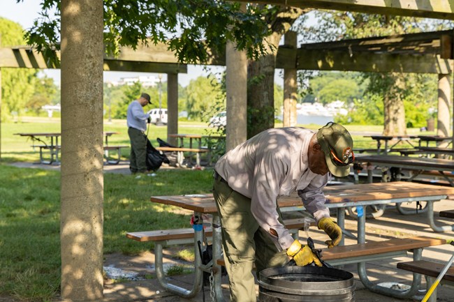 Two men in NPS uniforms pick up litter among a set of picnic benches in the shade.