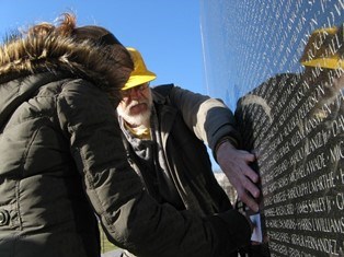 Two people stand in front of a memorial wall and read names