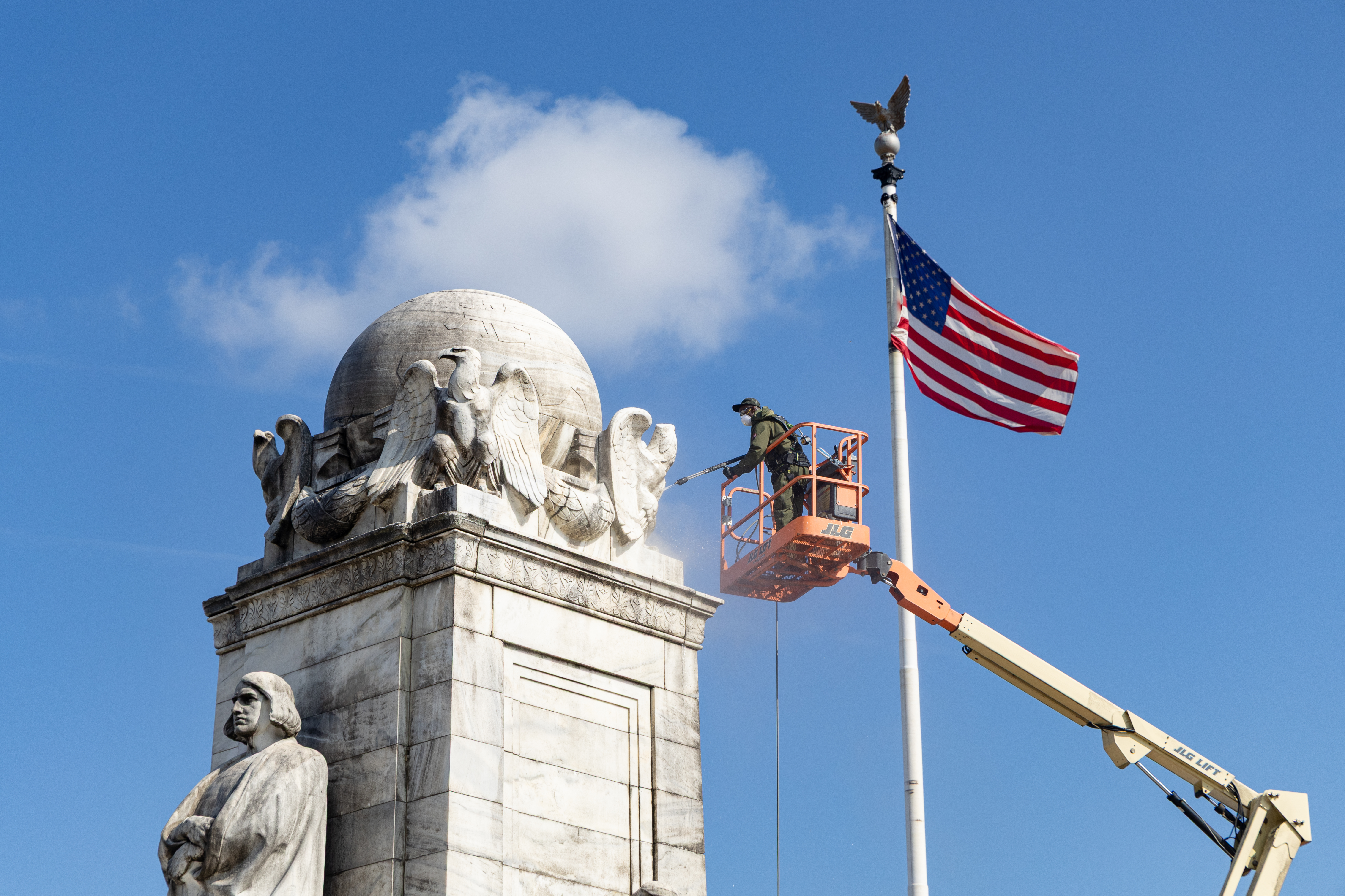 An NPS maintenance worker stands aloft in a cherry picker and uses a pressure washer to scrub a stone globe with eagles surrounding it, flanked by a large US flag.