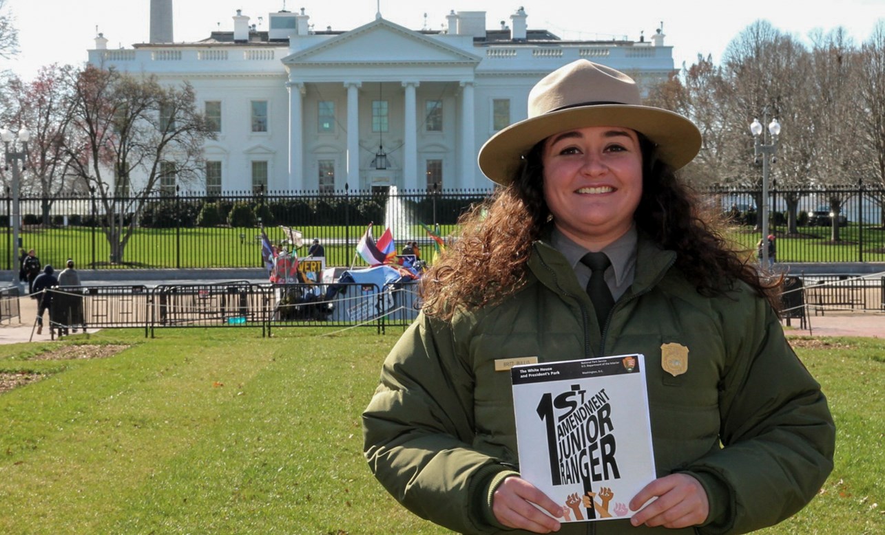 A woman in a green uniform jacket with a tan flat brimmed hat holds a booklets that reads "1st Amendment Junior Ranger" infront of the U.S. White House.