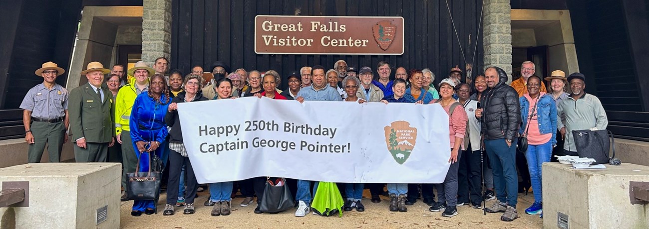 A large group of people holding a banner that reads "Hapy 250th Birthday Captain George Pointer!" under a sign that reads "Great Falls Visitor Center."