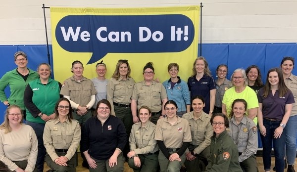 A group more than a dozen women smiling for a photo in front of a sign that reads "We Can Do It!"