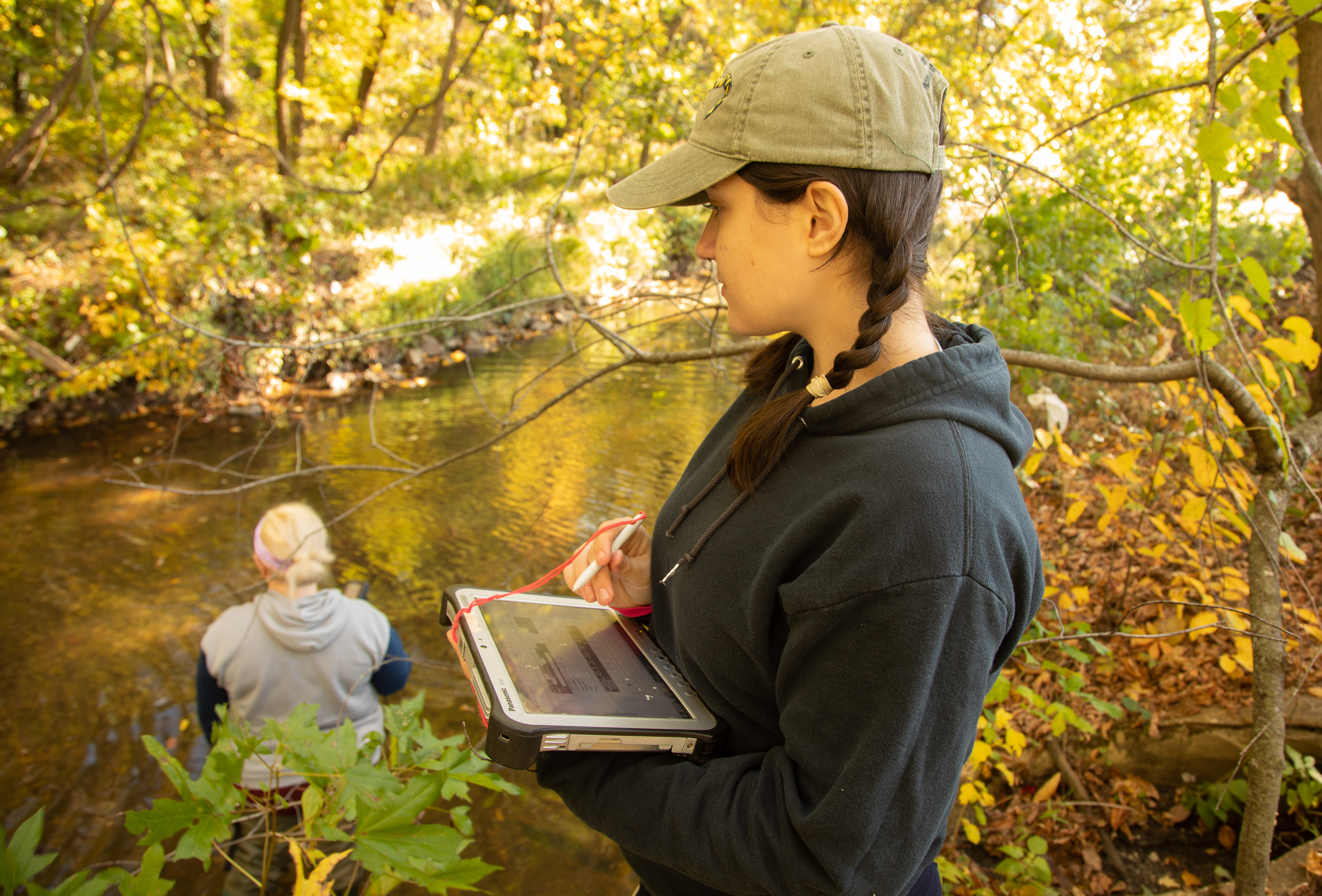 A person holding a tablet and stylus stands in a forest surrounded by branches and leaves. In the background, another person stands in a stream.