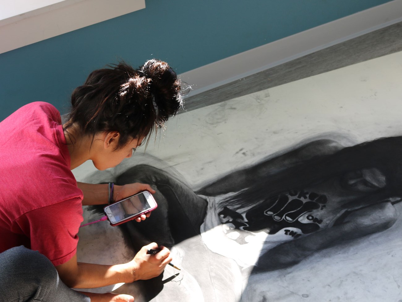 Artist Amy Araujo works on a large charcoal drawing, using her phone and a drawing tool. Sunlight highlights her focused process.