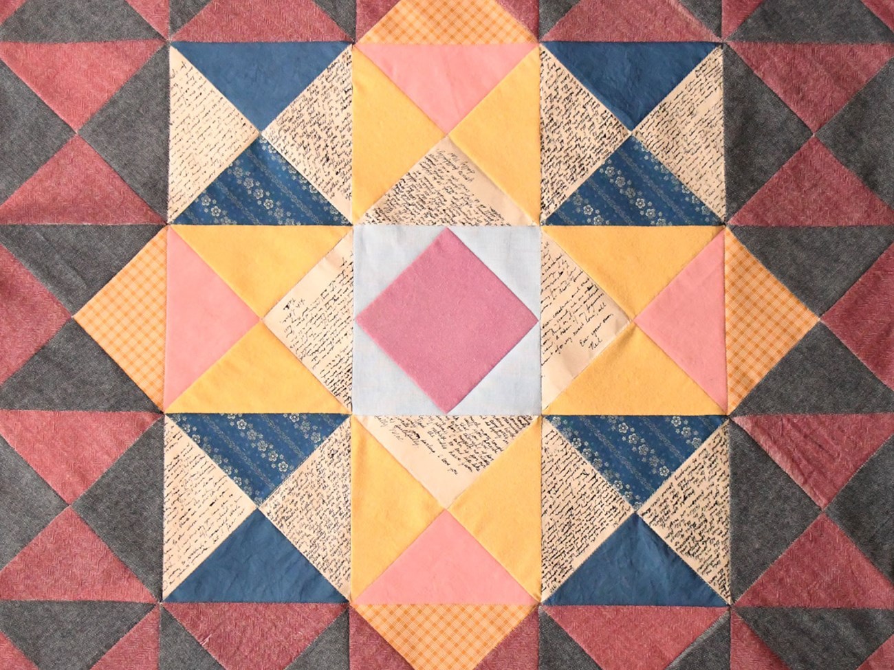 A geometric quilt with pink, blue, yellow, and gray triangles, some featuring text and floral patterns.