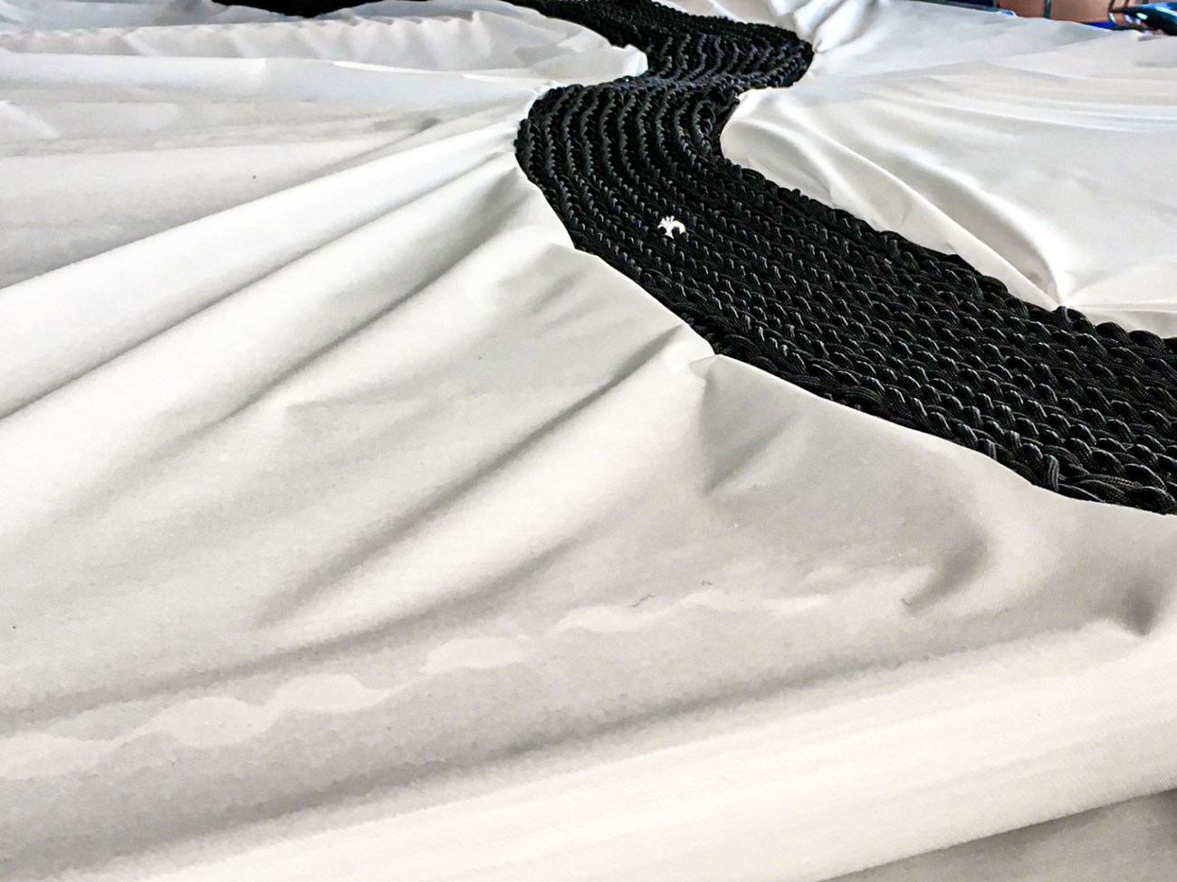 David Higgins' artwork featuring a hand-knit black road on white fabric, with strings below narrating the stories of displaced occupants through knotted data on their numbers, identities, and destinations.