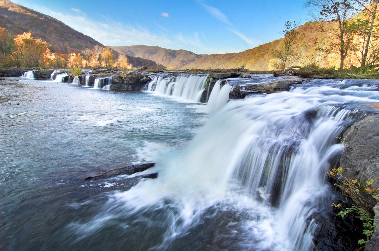 A wide shot of several waterfalls flowing into a large pool. Rolling hills and blue sky in background.