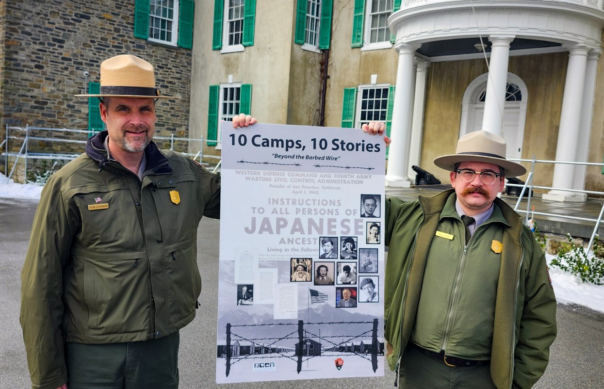 Two men in uniform hold up a sign that reads "10 Camps, 10 Stories: Behind the Barbed Wire" in of front of an old building.