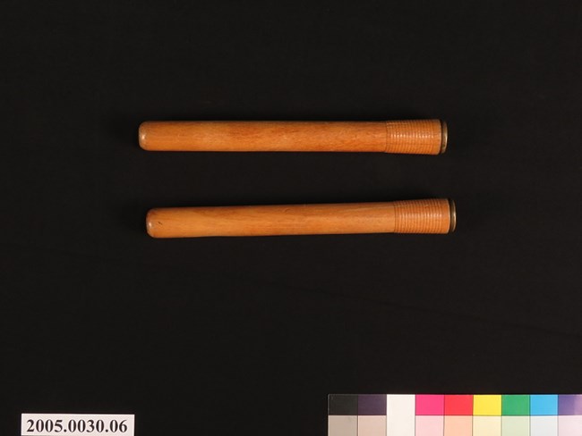 A set of two claves rest on a black surface. They are made of a light wood that is ribbed near the metallic end of each clave.