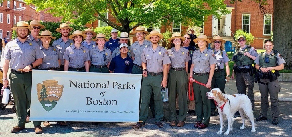 A large group of people stand together smiling in uniform with a sign that says, "National Parks of Boston." There is a dog on a leash with the group.