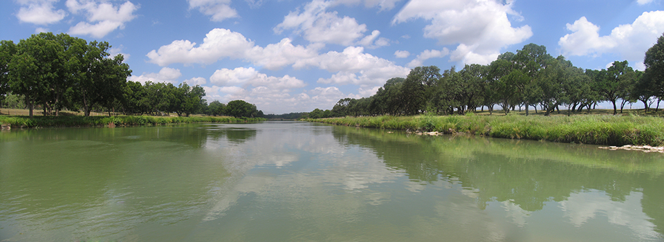 Panorama photo of the Pedernales River, with fluffy clouds in the sky and oak trees on both riverbanks.