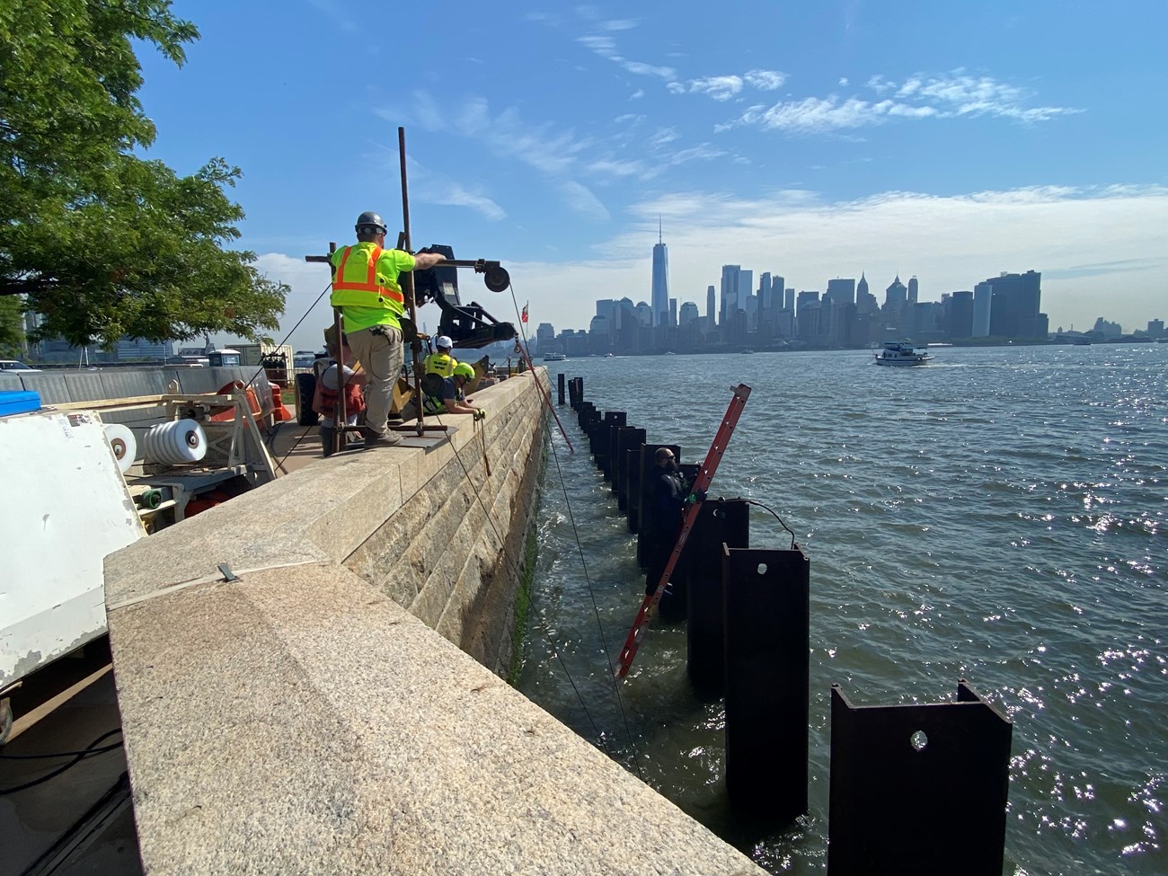 Construction workers operate machinery along a seawall with the city in the background.