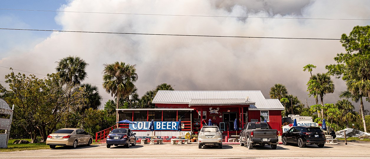 A red building with vehicles out front and a huge plume of smoke mushrooming behind and above.