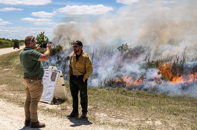 A man interviews a firefighter in Nomex while a fire burns in vegetation nearby.