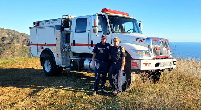 Two men stand in front of a fire engine, with the ocean in the background