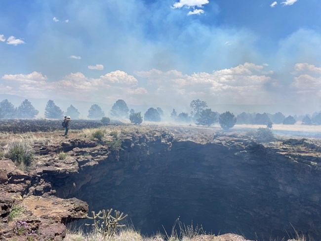 A firefighter stands near a large cave entrance with light smoke rising from the ground nearby