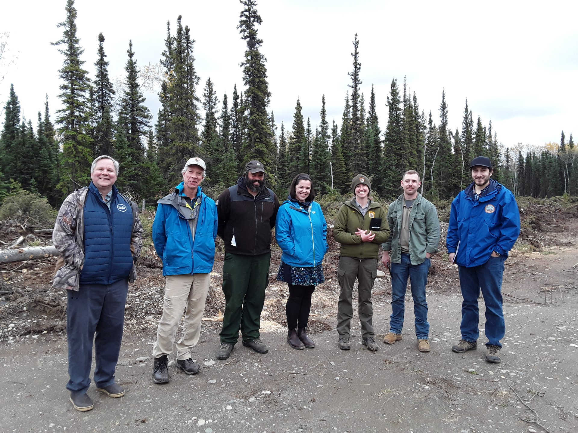 A group of 7 people smiles at the camera; trees in the background