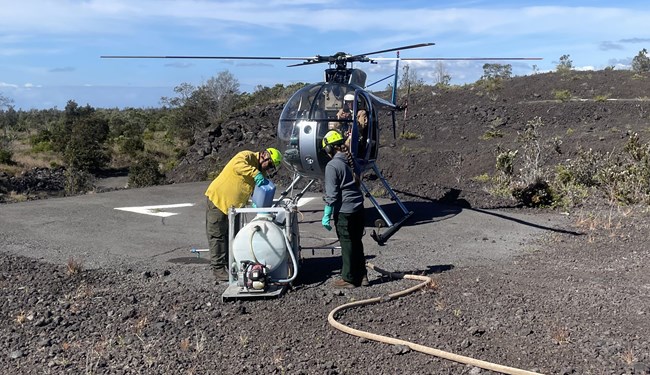 Two people in front of a helicopter; one is pouring something from a container into a larger apparatus