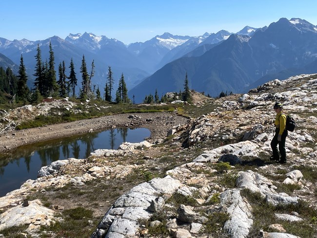 A person in wildland firefighter gear stands in a rocky area with mountains in the background