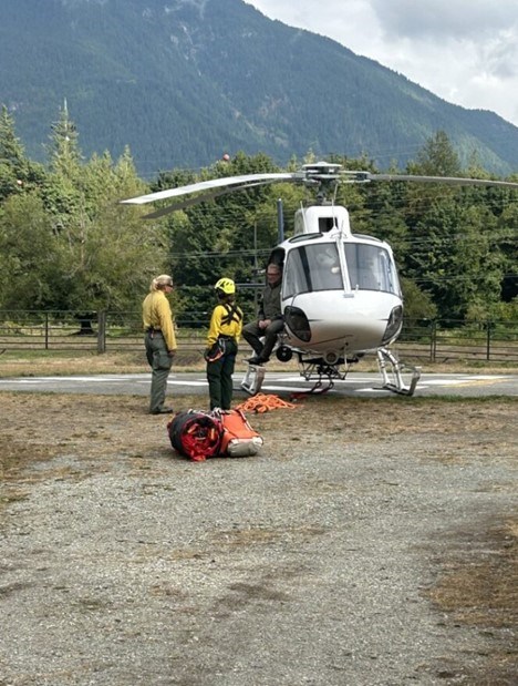 2 people in flight suits stand near a helicopter talking to a third person in the doorway of the helicopter
