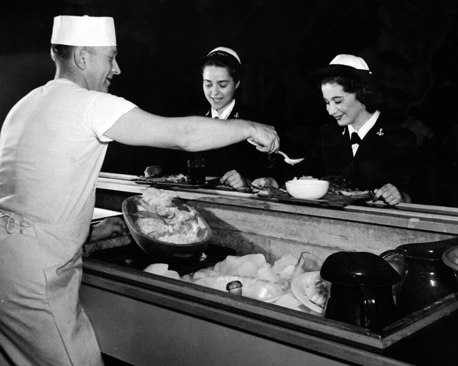 Black and white photo of two women in uniform being served food by a man behind a counter