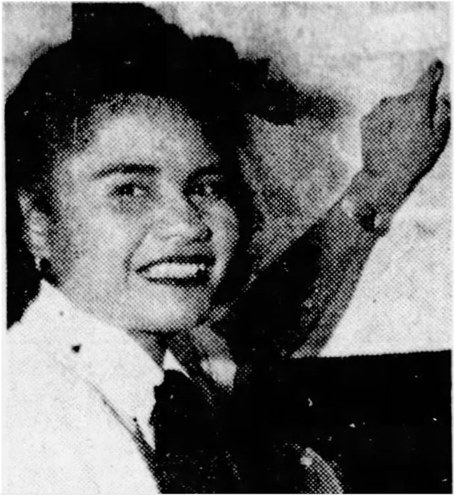 Grainy picture of a young woman with dark hair smiling at the camera. She has a bow in her hair and a uniform tie around her neck. She points to something behind her.