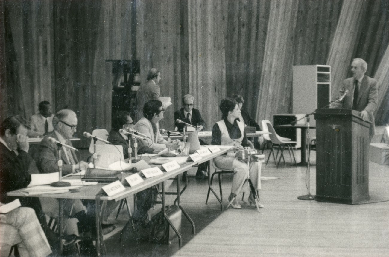 A man with white hair stands at a podium talking to a row of other mean, seated at microphones, while a female stenographer types.