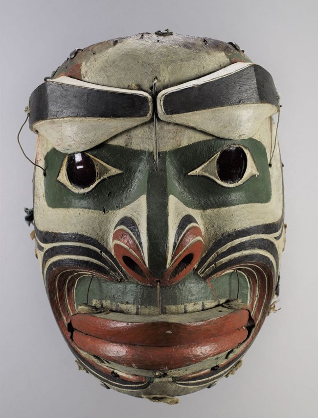 A painted mask of tan, brown, and red bands, green around trhe eyes, with brown eyebrows and a wide red mouth.