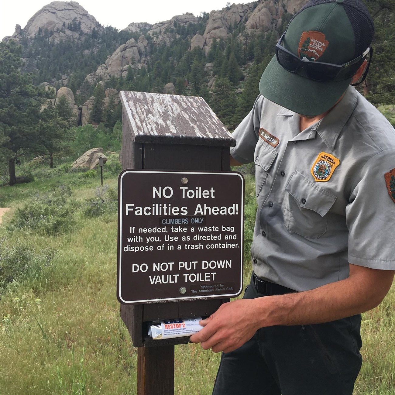 a ranger stands next to a WAG bag dispenser with sign that says "No toilet facilities ahead! If needed, take a waste bag with you. Use as directed and dispose of in trash container. Do not put down vault toilet."