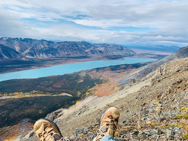 Hiking boots in the foreground. Vantage point view overlooking a blue lake surrounded by trees an mountains. Trees are green yellow, and red.