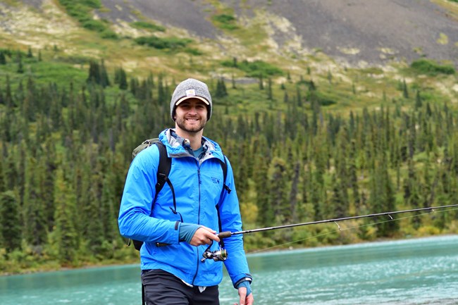 A man wearing a blue jack carrying a backpack stands in front of a turquoise-blue lake surrounded by trees.
