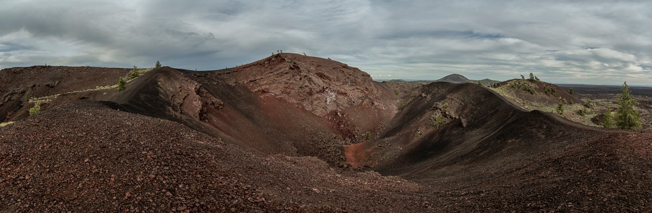 A wide shot of a crater, in a dark, volcanic landscape, dotted with sparse vegetation.