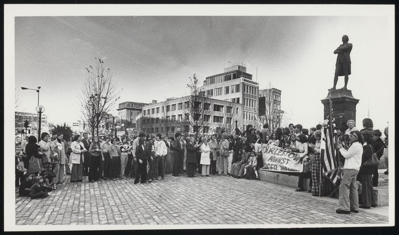 Black and white photo of a crowd standing next to the Samuel Adams statue holding signs that say Charlestown against forced busing