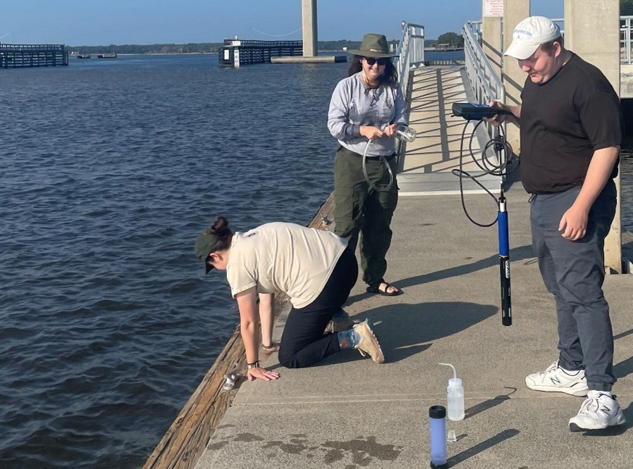 Three people on a dock. One kneeling on the edge of the water, the other two holding a sonde and water sample bottle.
