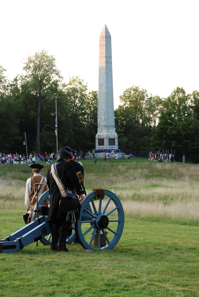 A tall stone obelisk is seen behind an 18th Century cannon crew.