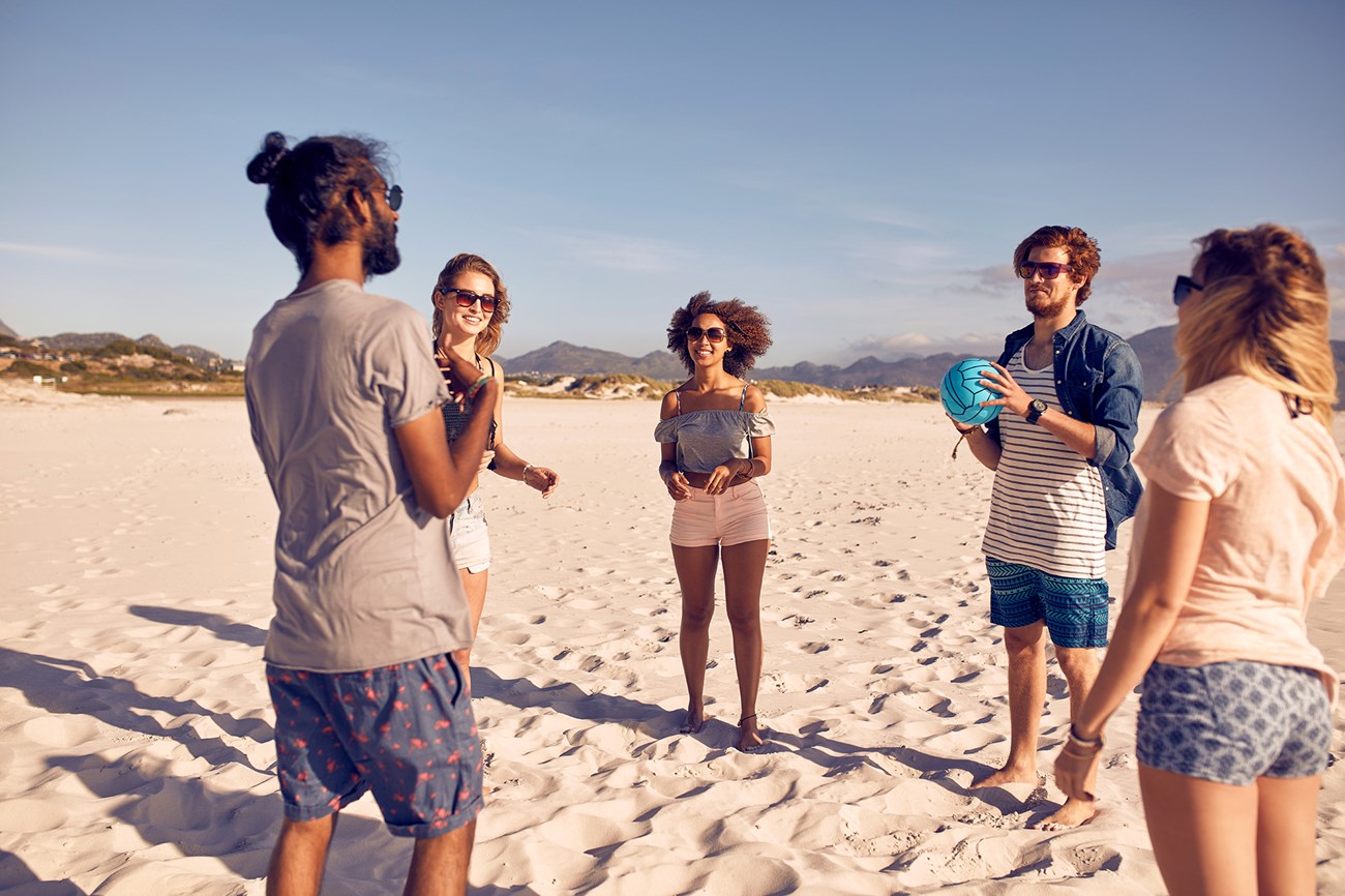 Five people stand on a beach, talking. One holds a volleyball.