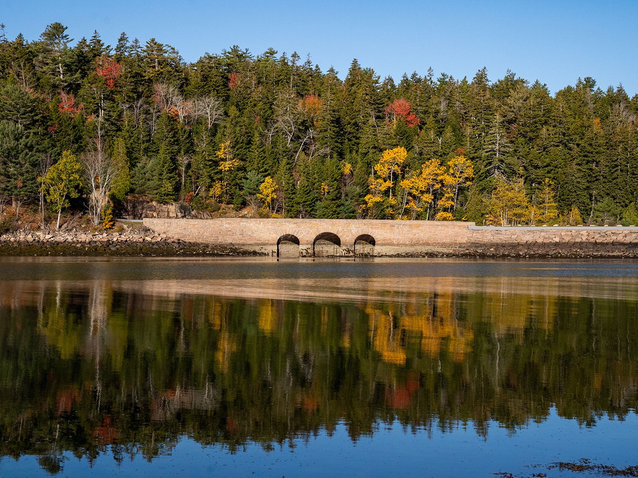 a triple arched stone bridge and fall foliage reflected in still water