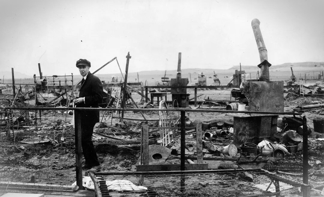 A man stands to the side of a field of broken stove pipes, twisted metal, and other debris from the destroyed miner’s tents.