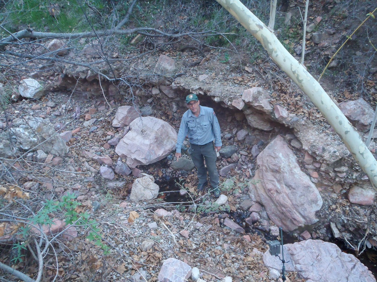 A man stands next to a small pool that drains in a narrow path down a slope. He points to the center of the pool for the camera.