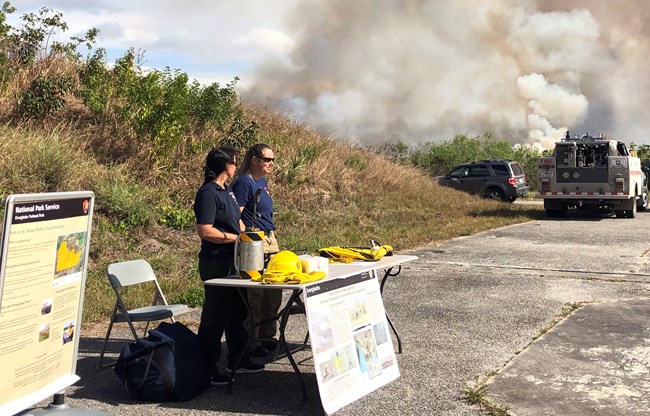 Two people stand behind a table with information in a parking lot with a fire engine and smoke billowing in the background.