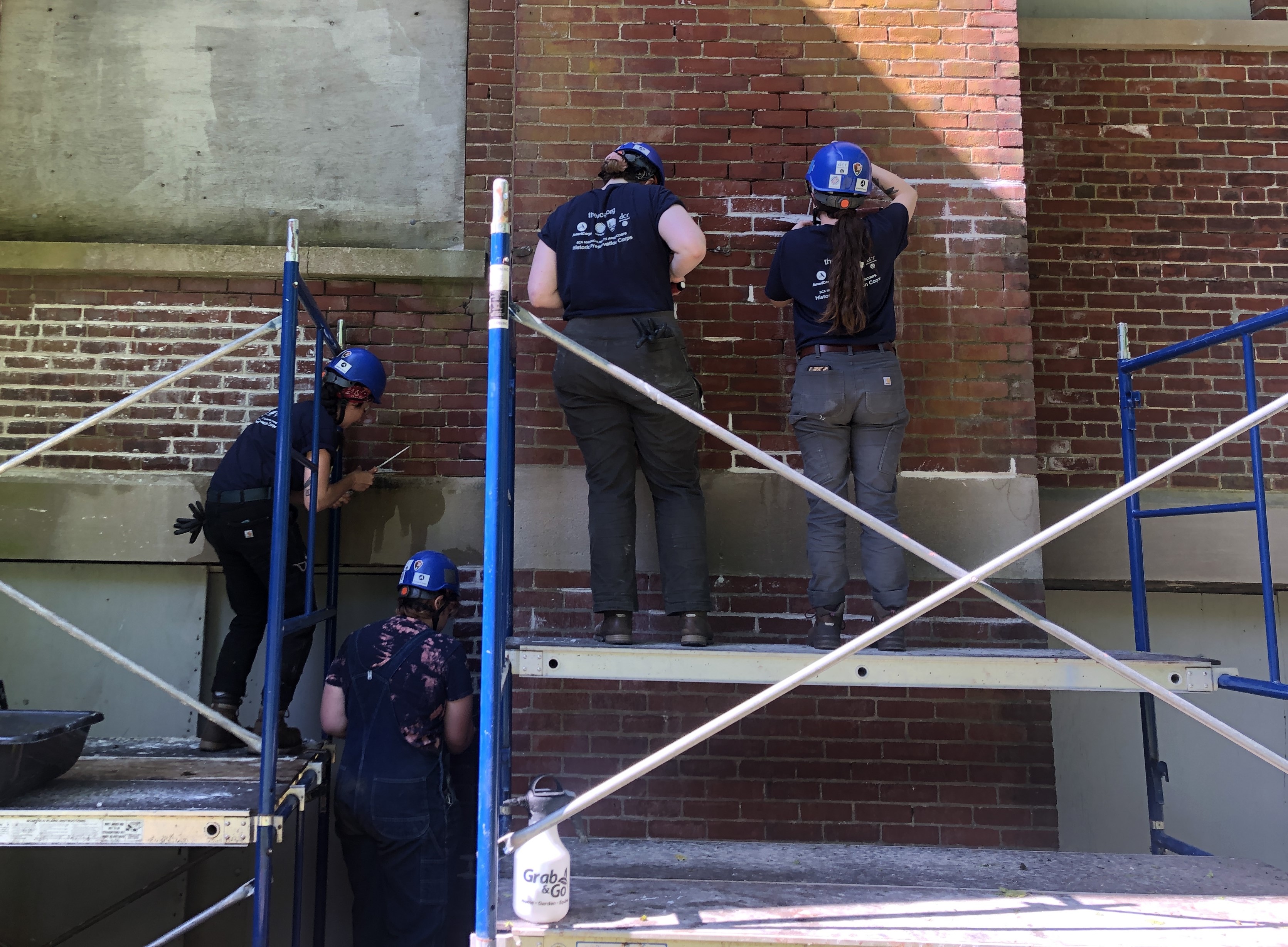 Four crew members wearing hard hats in front of an old brick wall. Three of them are on scaffolding, and one is standing on the ground below. The mortar in several parts of the wall is much brighter white than the mortar on the rest of the wall.