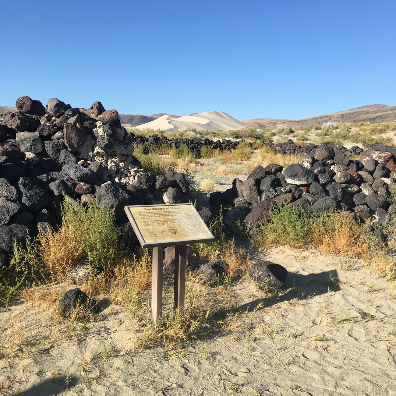 A sign stands in front of a ruined wall of rocks with distant sand dunes.