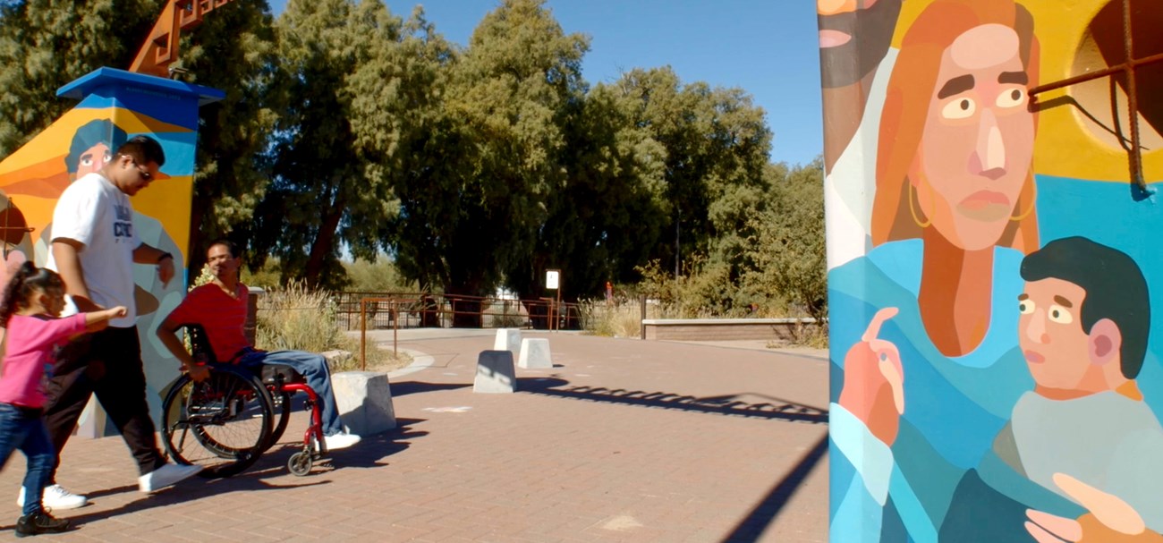 A person in a wheelchair, a man, and a girl walk along a brick pathway lined with murals of people.