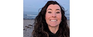 Fowler, with long, dark, wavy hair and a black hoodie, smiling at the camera at a beach.