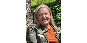 Headshot of Ziesler outside by a tree, smiling. She wears an orange shirt with a green outer layer with an NPS name badge affixed to it.