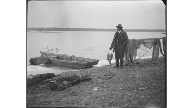 A black and white photograph of a man next to a rowboat and fishing net.