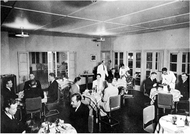 Several round tables with white table cloths, napkins, and good china fill a room. Many have 2 to 4 people (mostly men) sitting at them. They are being served by men in white uniforms. The lobby of the hotel is visible through an open door.