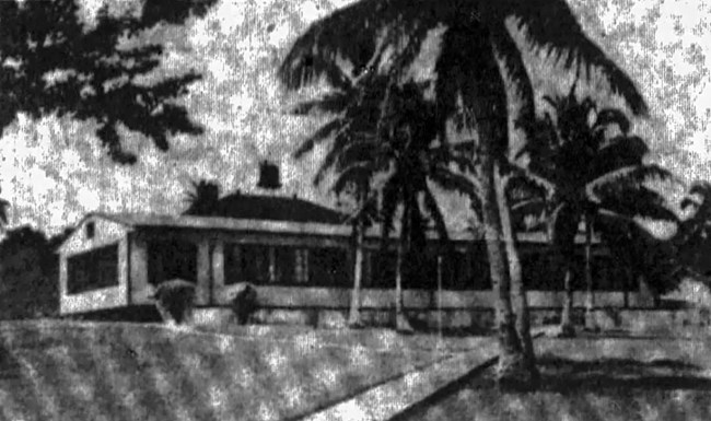 Black and white photo of the exterior of a one-story building with awnings over the windows. In the background are palm trees and hills.