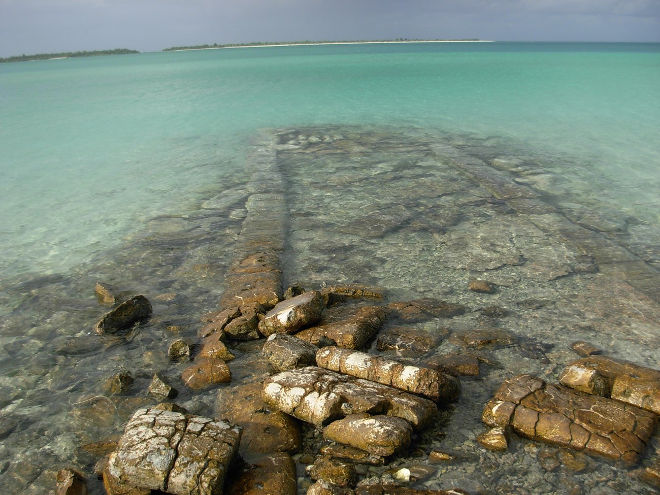 Color photograph of the ruins of a concrete structure extending into the bright turquoise lagoon of Wake Island. Across the lagoon are the other two islands that make up Wake.