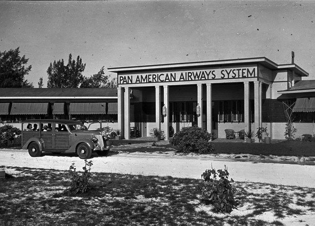 Black and white photo of a one story building. The center entrance has “Pan American Airways System” over it. Several people sit inside a vehicle driving on a shell-covered roadway. There are small plants and scrub grass.